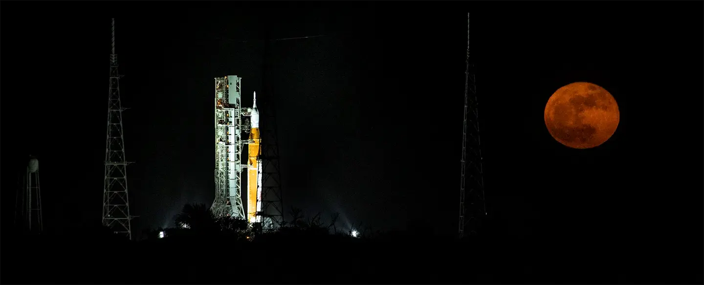 The Artemis Space Launch System on the launch pad with the moon floating behind in the night sky